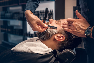 Barber shaving bearded male with a sharp razor. - obrazy, fototapety, plakaty