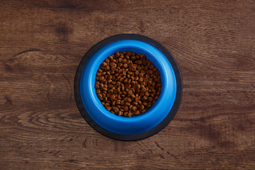 Bowl of dry kibble dog food. Healthy pets feed. Blue plate on wooden rustic background.