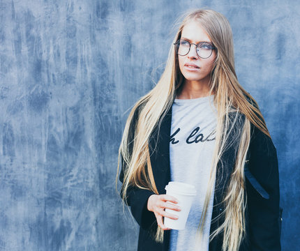Closeup portrait of blonde girl with long haircut with a paper cup of coffee. Hipster, insagram style. She wears black trendy dress, glasses, backpack. Outdoor.