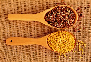 Jasmine red rice and peeled green peas in a wooden spoon on a sack background.