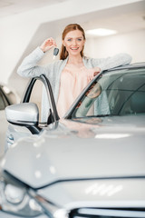 Happy woman holding car key and standing at car in dealership salon