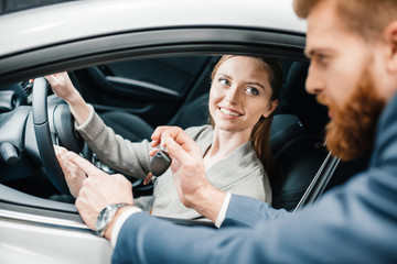 Bearded salesman giving car key to smiling young woman sitting in new car