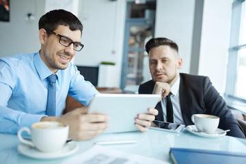 Waist-up portrait of young bearded employee holding digital tablet in hands and presenting his project to colleague, he listening to him with attention