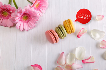 Macaroon cakes with pink rose petals and Gerbera flowers. Different types of macaron. I love you speech bubble. On white wooden rustic background.