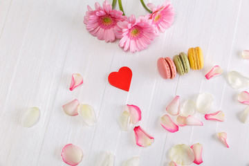 Macaroon cakes with red heart and Gerbera flowers. Different types of macaron. Colorful almond cookies. On white wooden rustic background.