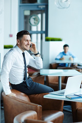 Joyful middle-aged businessman with stylish haircut talking to his colleague on smartphone while sitting in modern office lobby