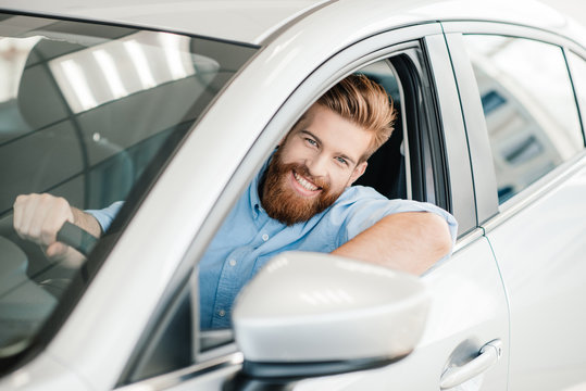 Smiling bearded young man sitting in new car and looking at camera