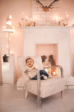 Cute baby girl sitting in retro chair in room. Wearing knitted sweater and denim pants. Looking at camera. Childhood.