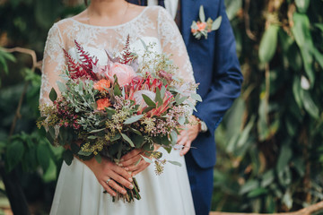 The groom in a suit and the bride in a white dress are standing on a green background, the bride is holding a wedding bouquet of flowers and greens with a ribbon