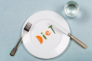 White plate with word diet made of colorful pieces of vegetables and glass of pure on blue tablecloth background. Flat lay. Top view. Concept of healthy eating and raw food dieting.
