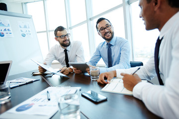 Multiethnic group of businessmen gathered in modern boardroom and discussing their joint project, one of them taking notes