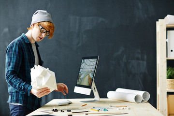 Young red haired architect holding house model and using modern computer standing at workplace with...