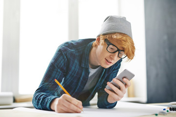 Fototapeta premium Portrait of young creative red haired man leaning on desk and using smartphone, browsing internet, while drawing with pencil on paper