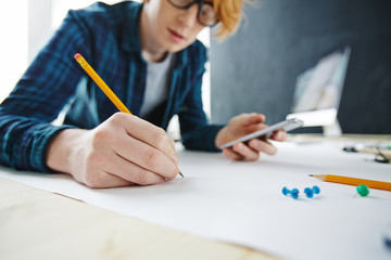 Portrait of young creative red haired man drawing and using smartphone at desk, focus on closeup hand holding pencil