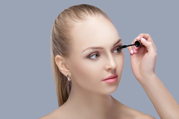 beautiful blond woman applying makeup on face on gray background. perfect makeup.  brush of mascara 