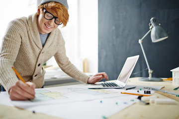 Obraz premium Portrait of smiling young red haired designer wearing glasses and beanie hat working in office: leaning on desk drawing floor plan and using laptop