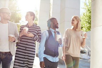 Multiethnic group of joyful financial managers walking together in city center and having small talk during coffee break, lens flare