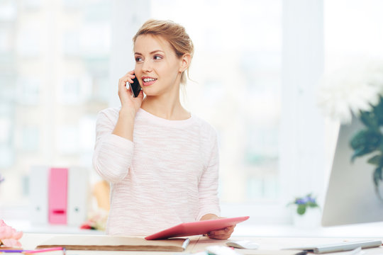 Pretty interior designer using smartphone to discuss project details with her client while standing in office against panoramic window