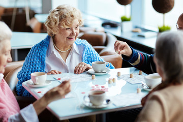 Group of four senior people gathered together in small cozy cafe, drinking fragrant tea and playing lotto with interest
