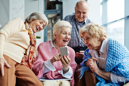 Four cheerful elderly people hanging out in spacious coffeehouse and looking through photos on modern smartphone