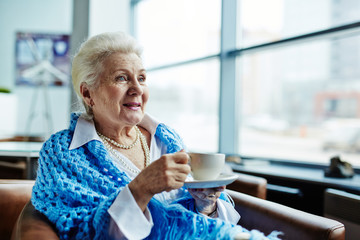 Pretty senior woman relaxing in cozy armchair and warming herself with cup of hot tea after long walk through winter city
