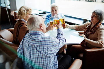 Group of joyful elderly people sitting in spacious cafe with panoramic windows and toasting with beer glasses