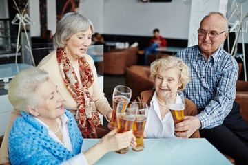 Having good time with friends in pub: group of joyful senior people sitting at table and clanging glasses of beer together