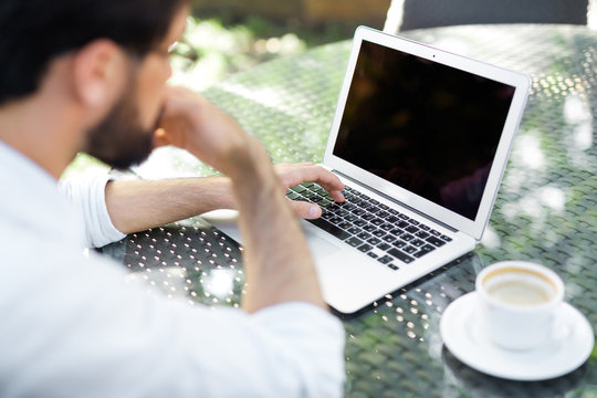 Bearded Financial Manager With Rolled Up Shirt Sleeves Searching Mistake In His Calculations On Laptop While Drinking Coffee In Outdoor Cafe, Over Shoulder View