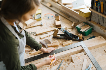 Profile view of confident young craftswoman marking measurement with help of steel framing square and pencil in workshop