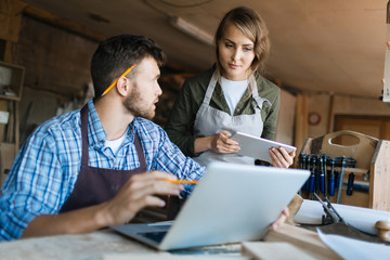 Pretty young carpenter and her bearded colleague brainstorming in workshop: he showing her...