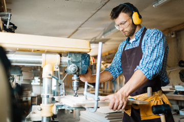 Bearded carpenter wearing apron and tool belt using drill press on wooden board while standing in workshop