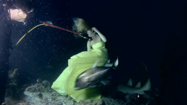 Scuba diver gives air to a young beautiful woman in a yellow dress posing underwater with a Tawny nurse sharks (Nebrius ferrugineus), a night shoot, the Indian Ocean, the Maldives
