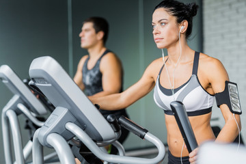 Fototapeta premium Portrait of beautiful sportive brunette woman exercising using elliptical machine and listening to music next to fit man during workout in modern gym