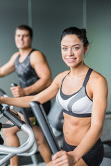 Fototapeta na wymiar Portrait of beautiful sportive brunette woman exercising using elliptical machine next to fit man, both smiling looking at camera working out in modern gym