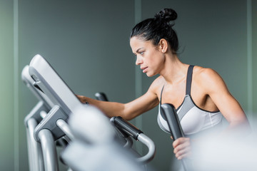 Fototapeta na wymiar Side view portrait of beautiful sportive woman exercising using elliptical machine during intense workout in modern gym