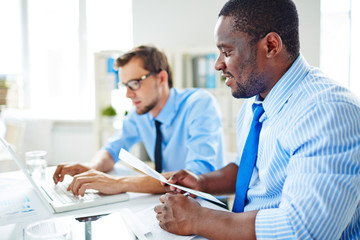 Obraz premium Middle-aged jurist in eyeglasses drawing up agreement on laptop while his Afro-American boss with document in hands keeping eye on him