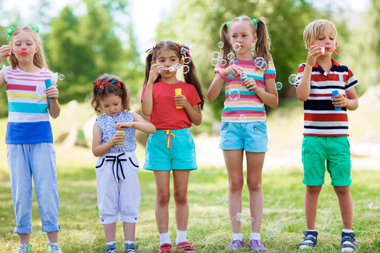 Children in summer wear standing in a row on green lawn and blowing soap bubbles