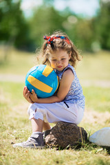 Portrait of cute little girl sitting alone on stone in park, holding ball and looking down pensively