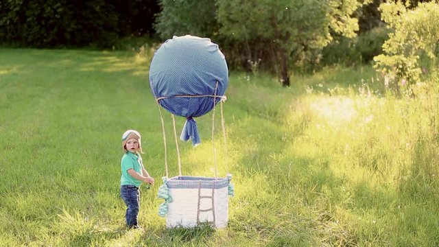 Beautiful funny little child wearing knitted pilot hat plays near handmade toy airballoon in nature landscape. Baby looks through binoculars. real time full hd video footage.
