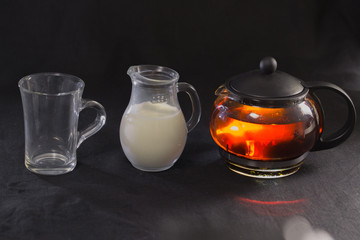 black tea in a transparent teapot glass tea Cup with milk in a small jug on black background.