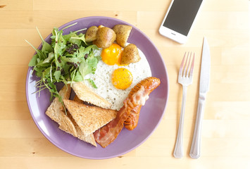 American breakfast with sunny side up eggs, sausages, potatoes, vegetables, toasted bread, soft sunlight. top view on the wooden table with fork, knife and a smartphone. Working day morning concept.