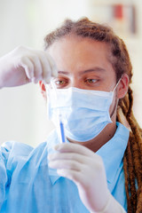 Young doctor with long dread locks posing for camera preparing syringe, wearing facial mask covering mouth, clinic in background, medical concept