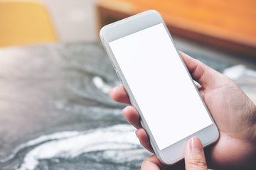 Mockup image of hand holding and touching white mobile phone with blank white screen on marble table in cafe