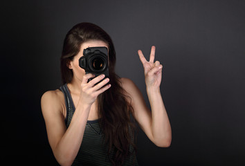 Young female photograph holding camera and makeing the photo and showing v-sign gesture on dark background with empty copy space. Closeup portrait