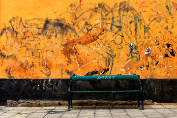 Orange wall covered in old graffiti and tags with a bench in front of it.