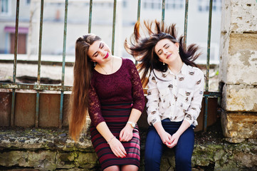Two young teenages girl sitting against iron fence and moving with they hair. Plump girl vs thin. Friends teen.