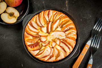 Apple pie in a portioned cast-iron frying pan, on a black stone table. With forks and apples Top...