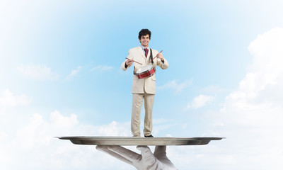 Young businessman on metal tray playing drums against blue sky background