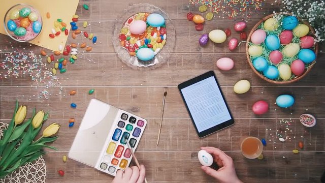 Man painting easter egg and digital tablet lies on table decorated with easter eggs Top view
