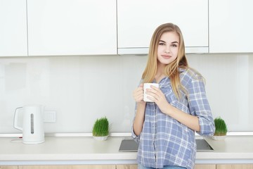 Young beautiful girl in a blue checkered shirt and jeans in the kitchen drinking tea or coffee with a white cup. Morning. home interior.
